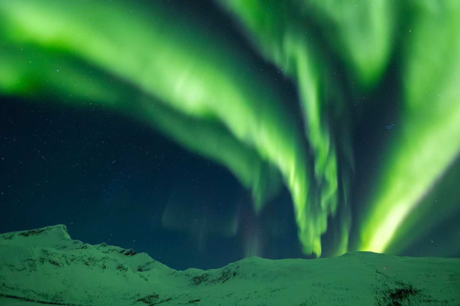 Bright green northern lights arching over snow-covered mountains under a starry night sky in Arctic Norway