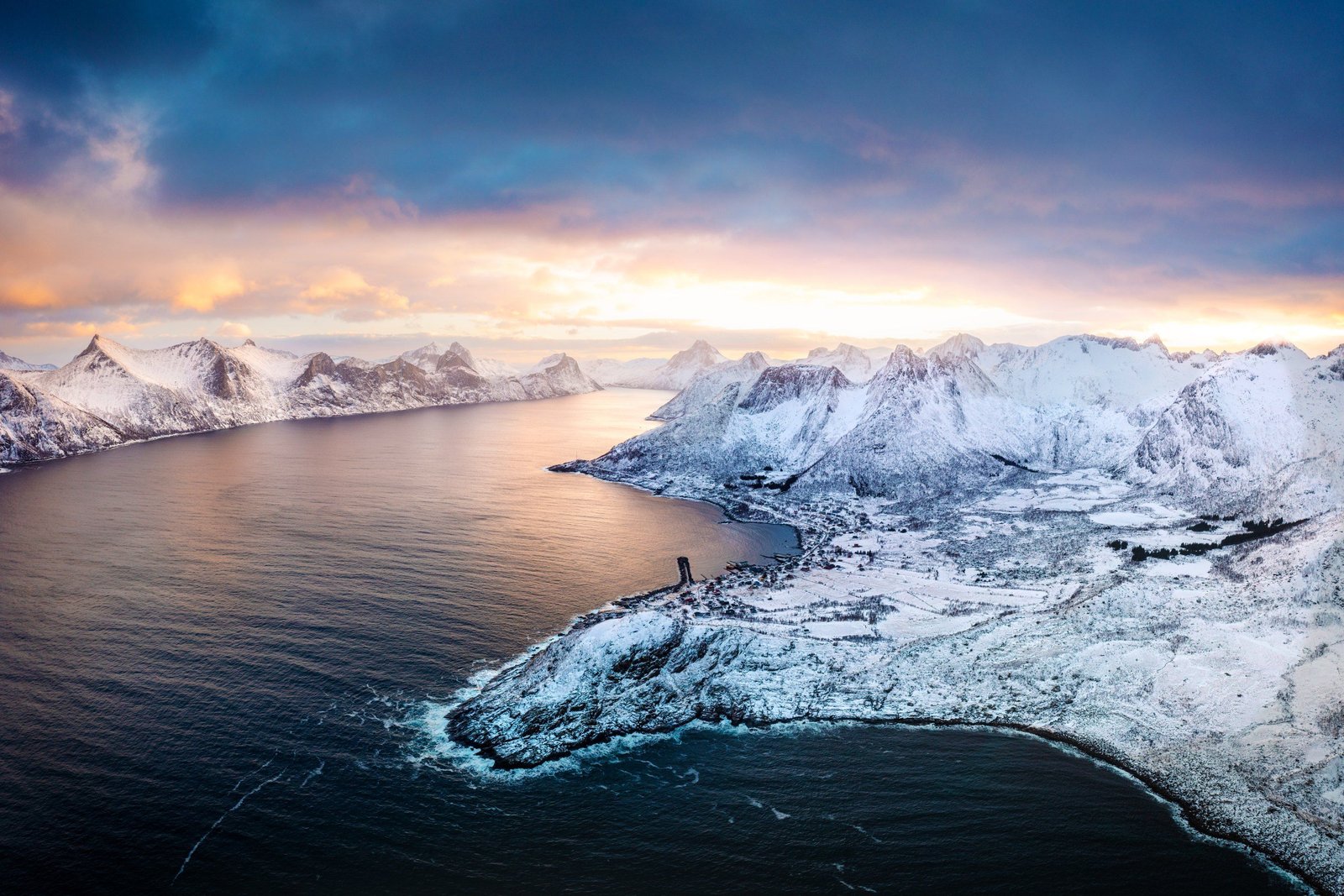 Aerial view of a snow-covered coastal village on a fjord, with white peaks under a pink and gold winter sky in Arctic Norway