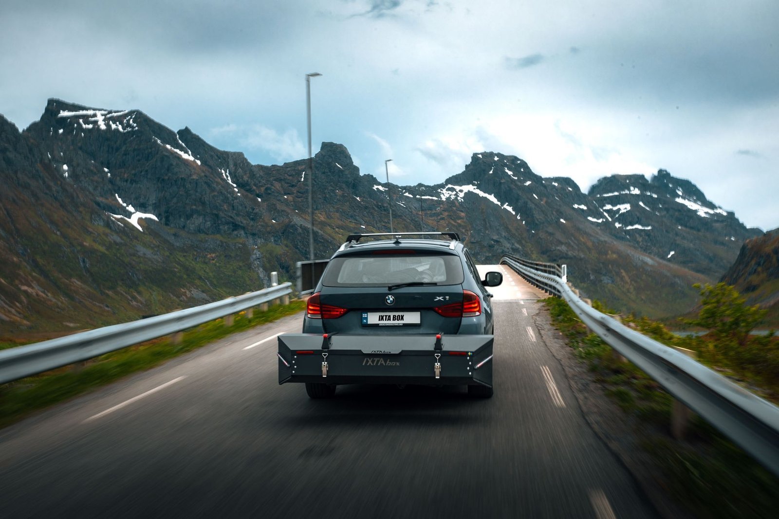 A car carrying an IXTA BOX drives a mountain road towards a bridge between snow-streaked peaks