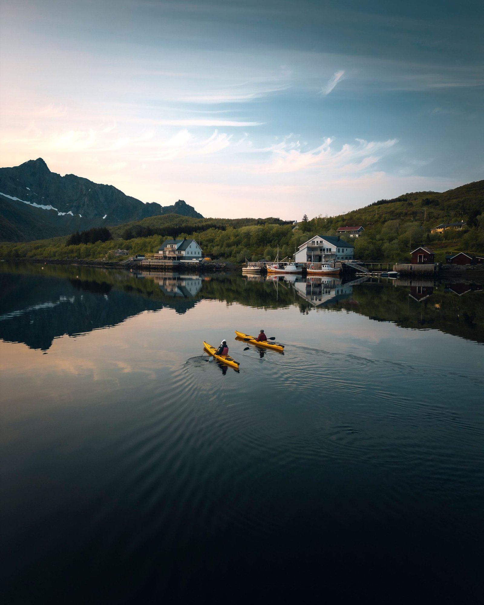 Two yellow kayaks glide across a mirror-calm bay towards white waterfront apartment houses below the mountains