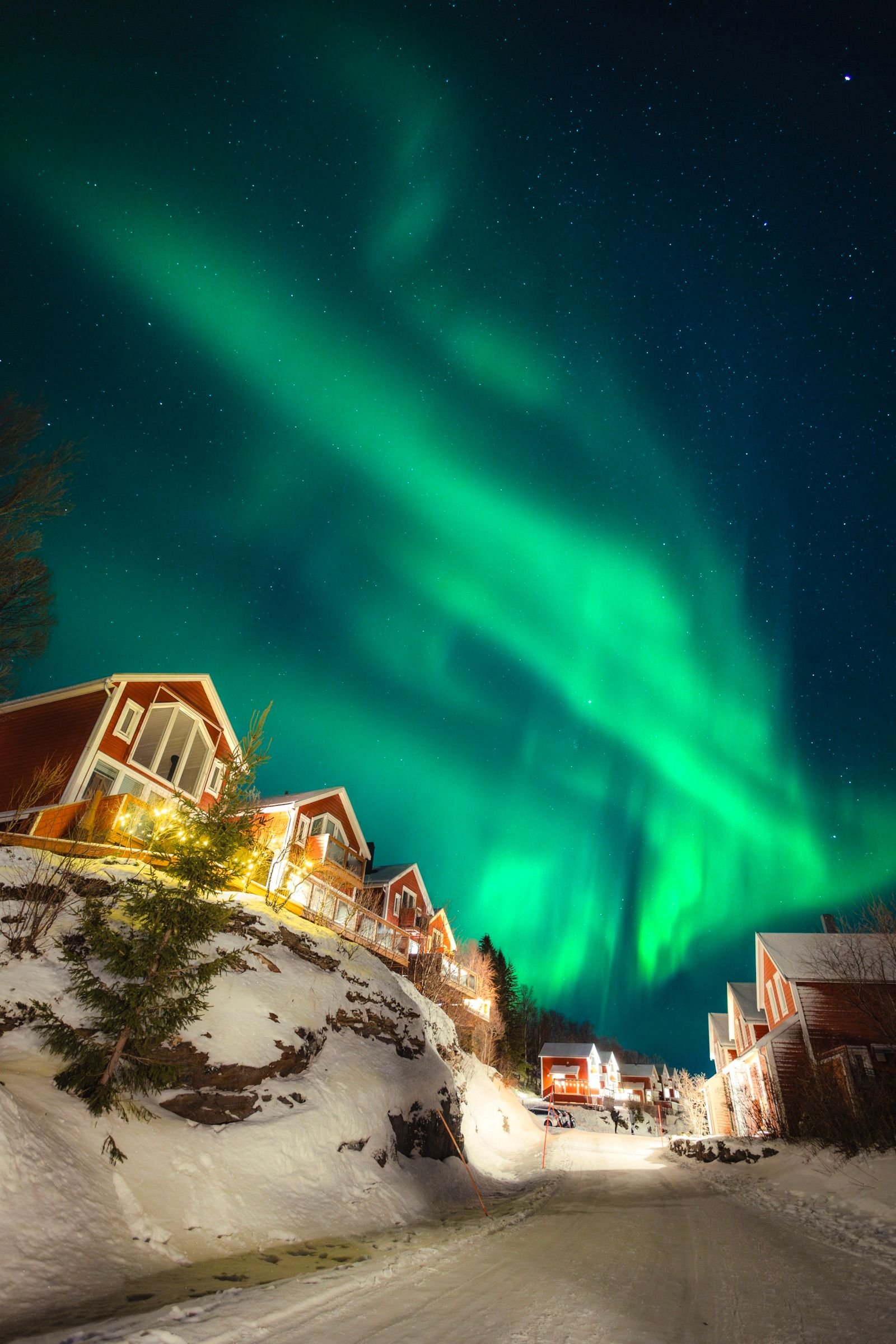 Green northern lights swirl over red resort houses lining a snowy road at night