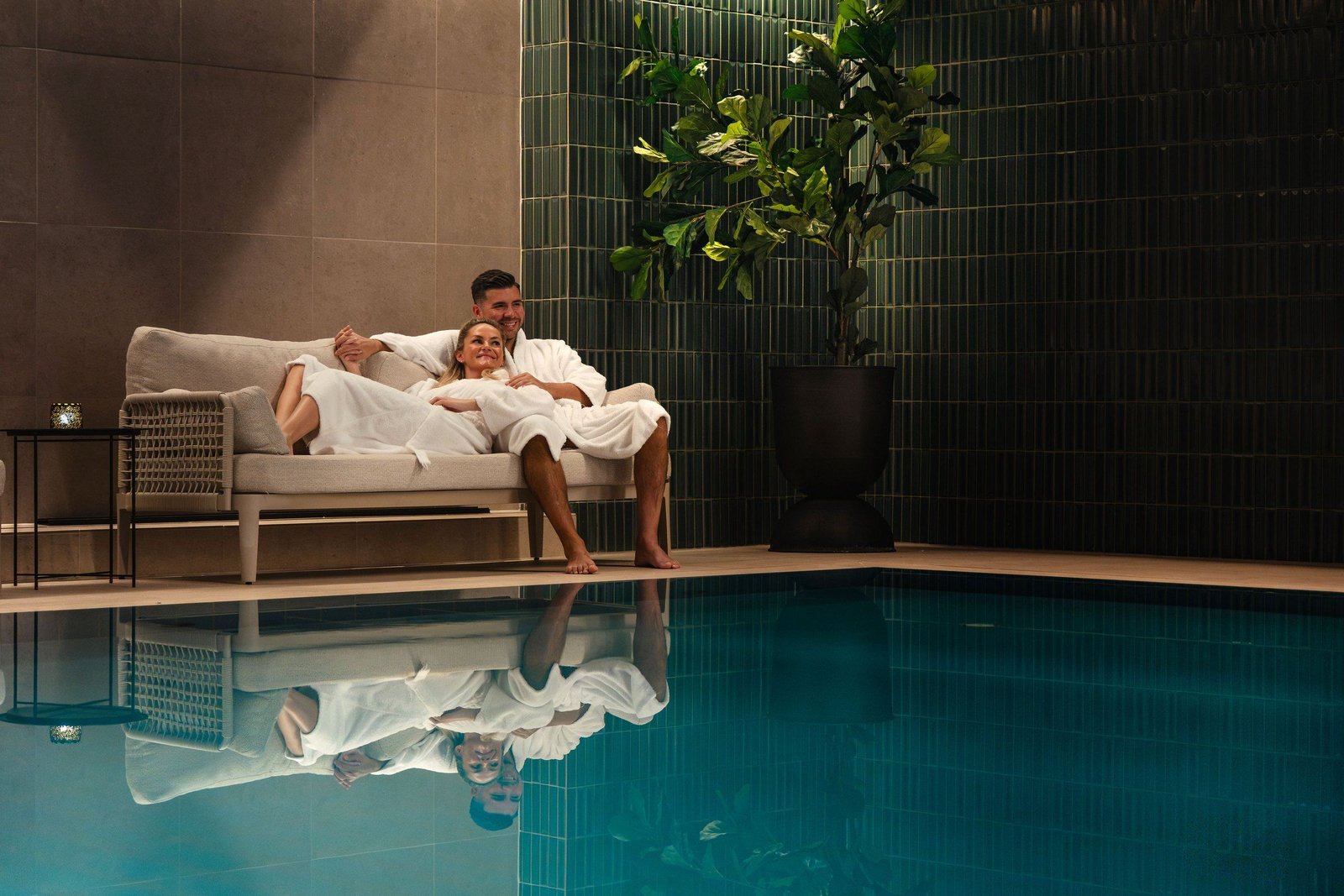 A couple in white bathrobes relax on a sofa beside an indoor pool with green tiled walls, reflected in the water
